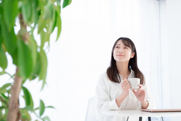 カップを手に持ちながら、穏やかな表情で窓の外を見つめる女性。気持ちを整え、前向きになっている様子