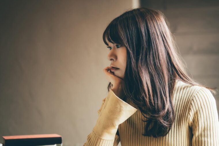 机に手をつきながら真剣な表情で考え込む女性。職場での人間関係に悩み、気持ちが沈んでいる様子